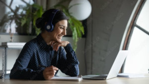 Smiling Indian female employee in wireless earphones talk on video call, have web conference with colleagues, happy ethnic woman worker in headset watch webinar on modern laptop at home